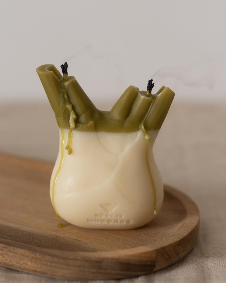 以下が含まれることがあります： 球根状の野菜の形をしたユニークなキャンドル。上部はオリーブグリーンで、アイボリー色のベースにワックスが垂れています。2本の消えた芯が見え、煙が立ち上っています。