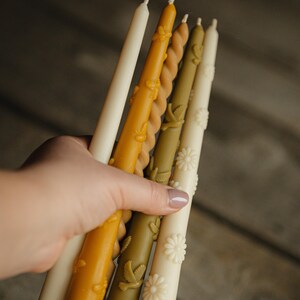 May include: A collection of five taper candles in varying shades of cream, yellow, olive green, and tan. The candles feature embossed details including small bees, birds, and daisy flowers, with one candle having a twisted spiral design.