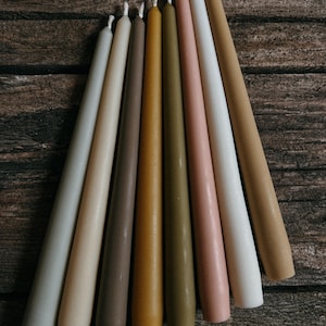 Puede incluir: Una colección de ocho velas cónicas dispuestas en forma de abanico, con una paleta de tonos tierra apagados que incluyen matices de gris, beige, marrón, amarillo mostaza, verde oliva, rosa empolvado y blanco roto, cada una con una mecha visible.