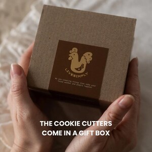 Op de afbeelding: Een bruine geschenkdoos met een decoratief etiket met een hanenillustratie en de tekst "LIVESIMPLY". De tekst "THE COOKIE CUTTERS COME IN A GIFT BOX" is ook zichtbaar.