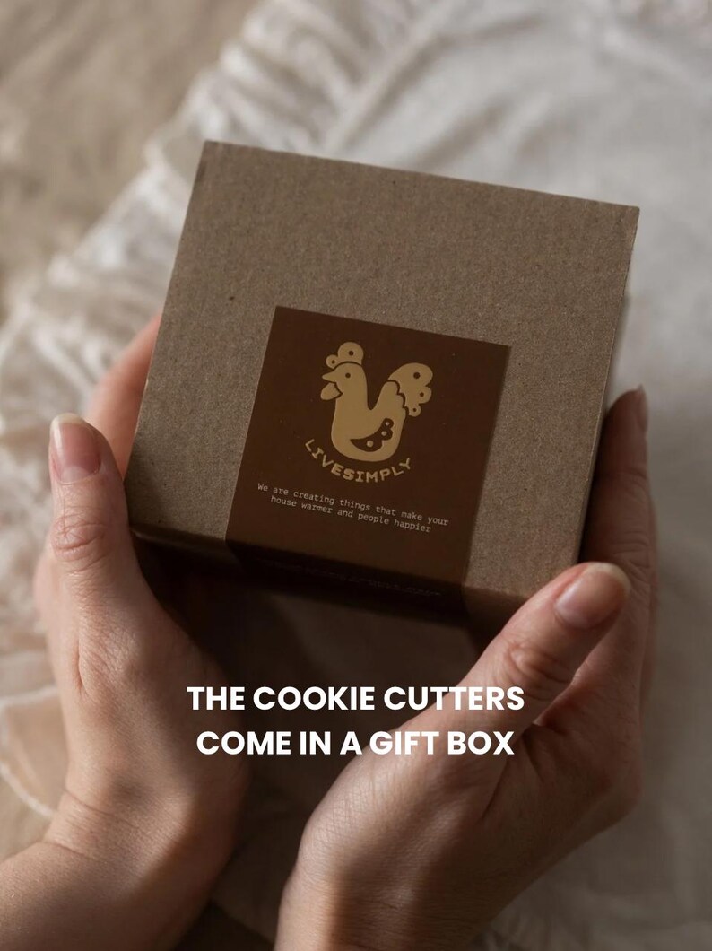 Op de afbeelding: Een bruine geschenkdoos met een decoratief etiket met een hanenillustratie en de tekst "LIVESIMPLY". De tekst "THE COOKIE CUTTERS COME IN A GIFT BOX" is ook zichtbaar.