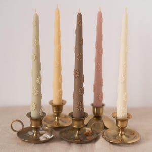 Könnte beinhalten: Fünf Stabkerzen in den Farben Salbeigrün, Hellgelb, Braun, Altrosa und Creme, die jeweils mit erhabenen Gänseblümchenmotiven verziert sind. Die Kerzen stehen aufrecht in einzelnen Messingkerzenhaltern mit kreisförmigen Sockeln und Griffen.