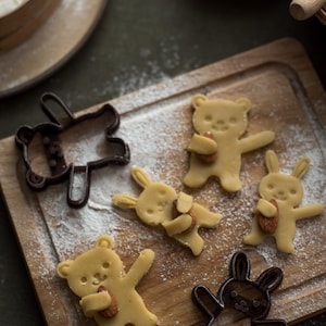 Pode incluir: Uma tábua de madeira com biscoitos em forma de urso e coelho e cortadores de biscoitos. Os biscoitos são amarelo claro e contêm amêndoas. A farinha é polvilhada sobre a tábua. Uma peneira e uma tigela de amêndoas estão no fundo.