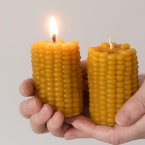 May include: Two yellow corn-shaped candles, one lit with a bright flame. The candles have a textured surface mimicking corn kernels. The unlit candle has a white wick. The candles are held in a person's hands.