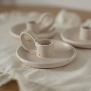 Peut inclure: Des bougeoirs en céramique blanc cassé avec une finition mouchetée. Chaque support a une base circulaire et une coupelle cylindrique pour la bougie. Un support a une poignée. Les supports sont sur un tissu blanc.