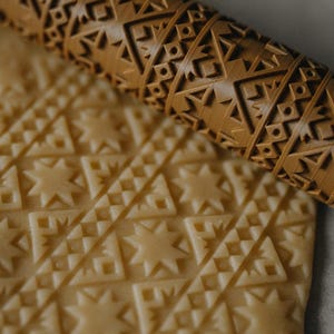 May include: A wooden rolling pin with a geometric pattern is shown partially rolled over dough. The rolling pin is light brown and features a repeating design of triangles, squares, and star shapes. The dough has the same embossed pattern.