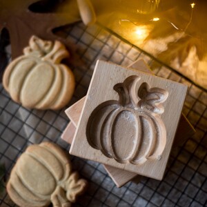 May include: A wooden pumpkin-shaped cookie mold with two baked cookies and a rolling pin. The mold is square and features a detailed pumpkin design. The cookies are light brown, and the scene is set on a wire rack.