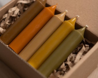 May include: A set of five taper candles in a cardboard box. The candles are in shades of brown, orange, and yellow. The box is filled with shredded paper for cushioning. The candles are a decorative item.