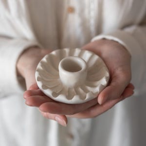 Puede incluir: Un pequeño candelabro de cerámica blanquecina con borde ondulado. El candelabro tiene una base circular y una abertura central cilíndrica para una vela. El diseño es sencillo y elegante, adecuado para la decoración del hogar.