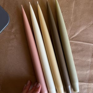 Pode incluir: Cinco velas cónicas dispostas em fila, com uma paleta de cores composta por rosa suave, branco sujo, amarelo claro, castanho escuro e verde sálvia. Cada vela tem um corpo cilíndrico liso que se estreita numa ponta no topo.