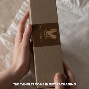 Op de afbeelding: Een bruine rechthoekige geschenkdoos met een bruine band met een logo en tekst. De doos wordt door twee handen vastgehouden. De tekst onderaan de afbeelding luidt: "DE KANDELAARS WORDEN GELEVERD IN GESCHENKVERPAKKING."