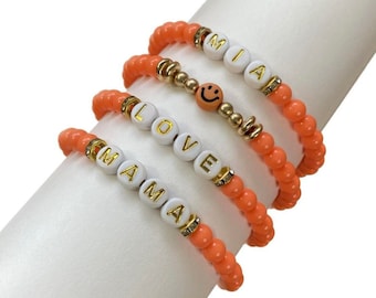 May include: Four beaded bracelets with the words "MIA", "LOVE", "MAMA", and a smiley face charm. The bracelets are all coral orange.