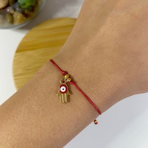 May include: Red string bracelet with a gold charm featuring a hamsa hand with an evil eye in the centre.