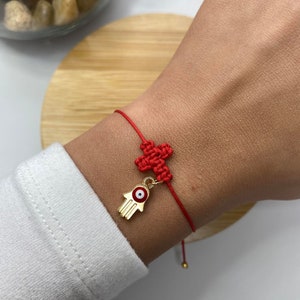 May include: Red string bracelet with a gold hamsa hand charm featuring a blue evil eye. The bracelet is tied with a knot and has a small gold bead at the end.