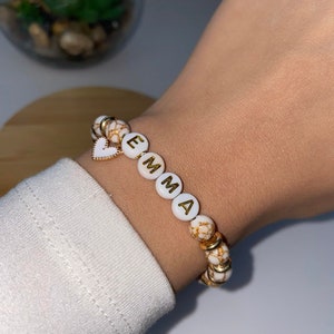 Peut inclure: Bracelet en perles blanches avec inscription dorée "EMMA" et un petit charm en forme de cœur doré.