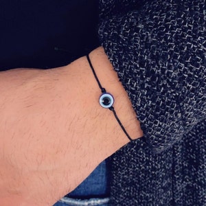 May include: A black string bracelet with a blue and white evil eye charm. The charm is circular, with a dark blue border, a white centre, and a black pupil. The bracelet is worn on a wrist.