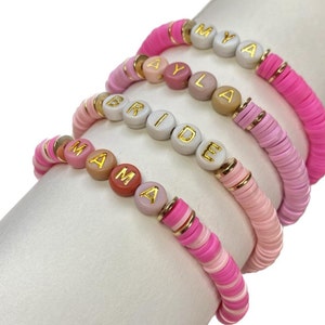 May include: Four pink and white beaded bracelets with gold lettering spelling out the names "MYA", "AYLA", "BRIDE", and "MAMA".