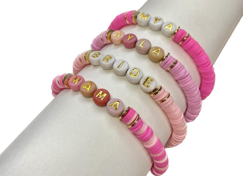 May include: Four pink and white beaded bracelets with gold lettering spelling out the names "MYA", "AYLA", "BRIDE", and "MAMA".