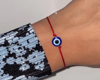 May include: A red string bracelet with a blue evil eye charm. The bracelet is tied around a person's wrist.