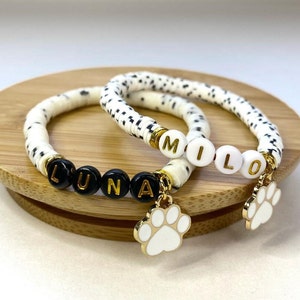 Peut inclure: Deux bracelets en perles blanches et noires avec des pendentifs en forme de patte d'or. Les bracelets épellent les noms "Luna" et "Milo" en perles noires.