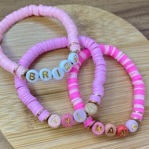 May include: Three pink and white beaded bracelets with gold accents. The bracelets spell out the words "Bride", "Ayla", and "Mama".