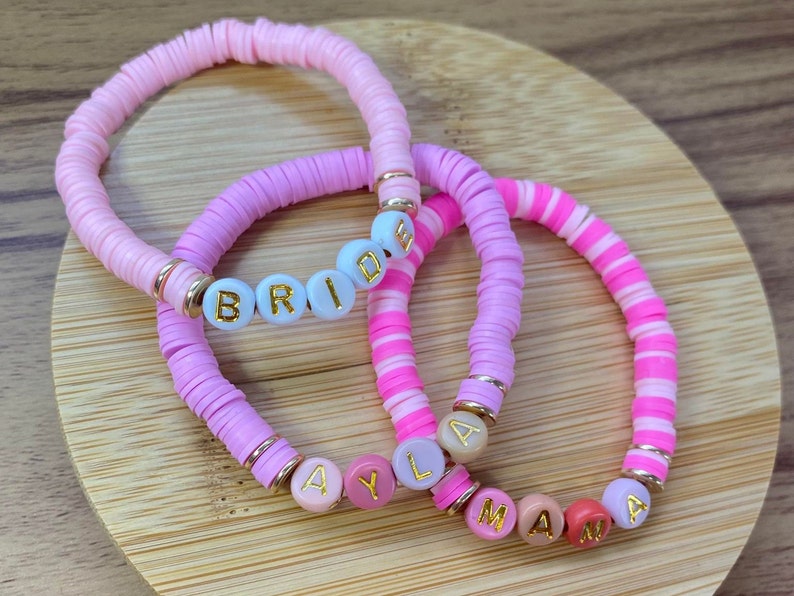 May include: Three pink and white beaded bracelets with gold accents. The bracelets spell out the words "Bride", "Ayla", and "Mama".
