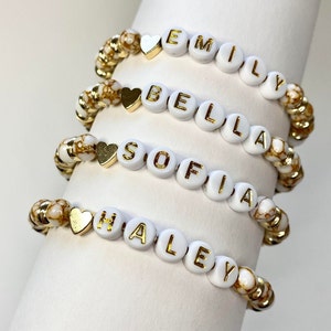 Peut inclure: Quatre bracelets personnalisés avec des perles de lettres blanches épelant les noms : Emily, Bella, Sofia et Haley. Chaque bracelet a des breloques en forme de cœur dorées et des perles d'accent marbrées dorées et blanches. Les bracelets sont exposés sur une surface blanche.
