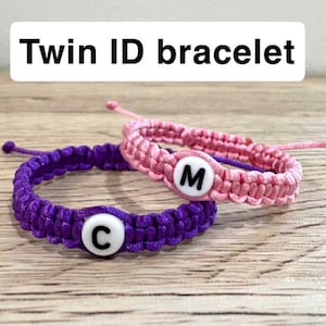 May include: Two handmade ID bracelets. One is purple with a white letter "C", the other is pink with a white letter "M". The bracelets are made of braided cord. The text "Twin ID bracelet" is in a white rectangle.