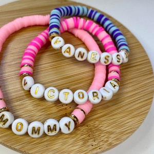 May include: Three pink and white beaded bracelets with gold lettering spelling out the names "Mommy", "Victoria", and "Vanessa".