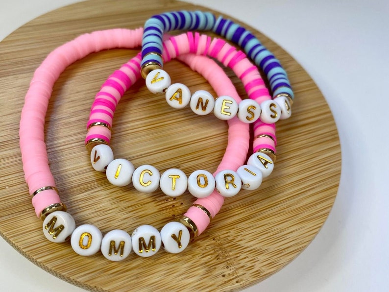 May include: Three pink and white beaded bracelets with gold lettering spelling out the names "Mommy", "Victoria", and "Vanessa".