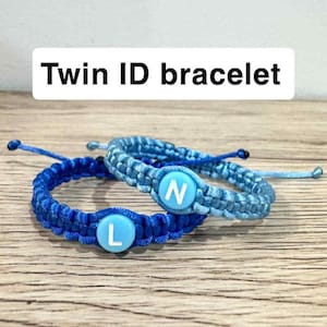 May include: Two blue braided bracelets with white lettered circular charms. One bracelet is a darker blue, the other is a lighter blue. The text "Twin ID bracelet" is in a white rectangle above the bracelets.