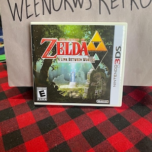 May include: The Legend of Zelda: A Link Between Worlds video game cartridge for the Nintendo 3DS. The game cover features a green background with a sword and a character in the middle. The Nintendo 3DS logo is on the right side of the cover.