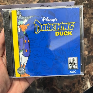 May include: A blue and yellow video game cartridge case for the TurboGrafx-16 console. The case features a cartoon image of Darkwing Duck, a Disney character, wearing a blue suit and a black hat. The text "Disney's Darkwing Duck" is printed in yellow on the case, along with the TurboGrafx-16 logo and the NEC logo.
