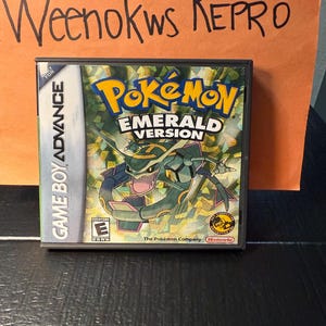 Puede incluir: Un cartucho de juego de Game Boy Advance para Pokémon Versión Esmeralda. La caja del juego presenta el logotipo de Pokémon en amarillo y la imagen de una criatura con forma de dragón verde y azul. El texto "GAME BOY ADVANCE" es visible en el lateral.