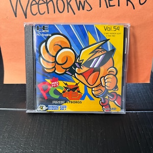 Op de afbeelding: Een PC Engine game case met een cartoon cyborg karakter met zonnebril op de cover. De illustratie is levendig met blauwe, gele en oranje tinten. Tekst bevat "PUNKIC CYBORGS" en "HUDSON SOFT".