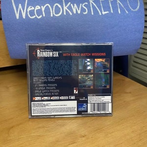 May include: Back of a Tom Clancy's Rainbow Six video game case with a blue background. The game features Eagle Watch missions and special forces action. The case is rated Teen 13+ for animated blood and gore and animated violence.