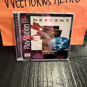 May include: A PlayStation game case for "Descent" featuring a futuristic spaceship design in red and purple. The case has the PlayStation logo and the text "Sensory Overload in 360 3D".