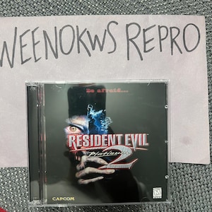 Puede incluir: Una caja de CD negra con una imagen roja y azul de una cara de zombi. El texto "Resident Evil Platinum 2" está impreso en blanco en la caja. La clasificación PEGI "M" para Mayores de 18 años está en la esquina inferior derecha.