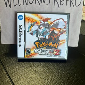 Könnte beinhalten: Eine Nintendo DS-Spielkassette für Pokemon White Version 2. Das Spielgehäuse ist schwarz mit einem roten und weißen Pokemon-Logo. Das Spiel zeigt einen weißen und blauen Pokemon-Charakter auf der Vorderseite.