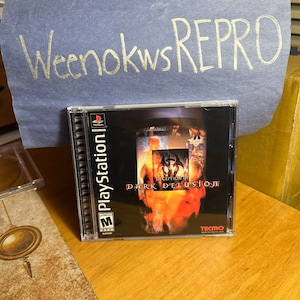 Puede incluir: Una caja de juego de PlayStation para Deception III: Dark Delusion. La caja es negra con una imagen roja y naranja de una mujer con una espada. El juego está clasificado como M para Maduros por la ESRB. El juego es publicado por Tecmo.