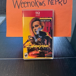 Może przedstawiać: Etui na grę wideo do gry na Nintendo Switch, Cyberpunk 2077: Ultimate Edition, Phantom Liberty. Grafika na okładce przedstawia kobietę z rudymi włosami i inne postacie. Etui znajduje się w przezroczystym plastikowym etui z pomarańczowym papierem nad nim z napisem "Weenokws REPRO".