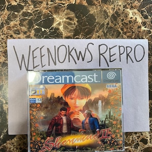 May include: A Dreamcast video game case with a blue and white background. The game is called "Shenmue II" and features a woman with a yellow hat and two men. The game is rated "12" by the BBFC.