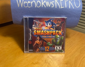 May include: A Sega Dreamcast game case with a blue background and the text "SEGA SMASHPACK VOLUME 1" in white. The case features a variety of characters from different games, including Sonic the Hedgehog, a green alien, and a muscular man. The case also includes the text "GREAT 12 GAMES" and an ESRB rating of "E-T for Everyone".