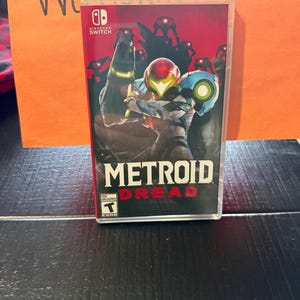 May include: A Nintendo Switch game case for "Metroid Dread" featuring a red and black background with a character in a suit holding a weapon. The game title is in large white letters with the word "Dread" in red.