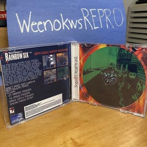 May include: A Sega Dreamcast game case for Tom Clancy's Rainbow Six. The front of the case shows a dark, urban scene with a person in the foreground. The back of the case has a white background with black text and a picture of the game's logo. The text on the back of the case reads "With Eagle Watch Missions".