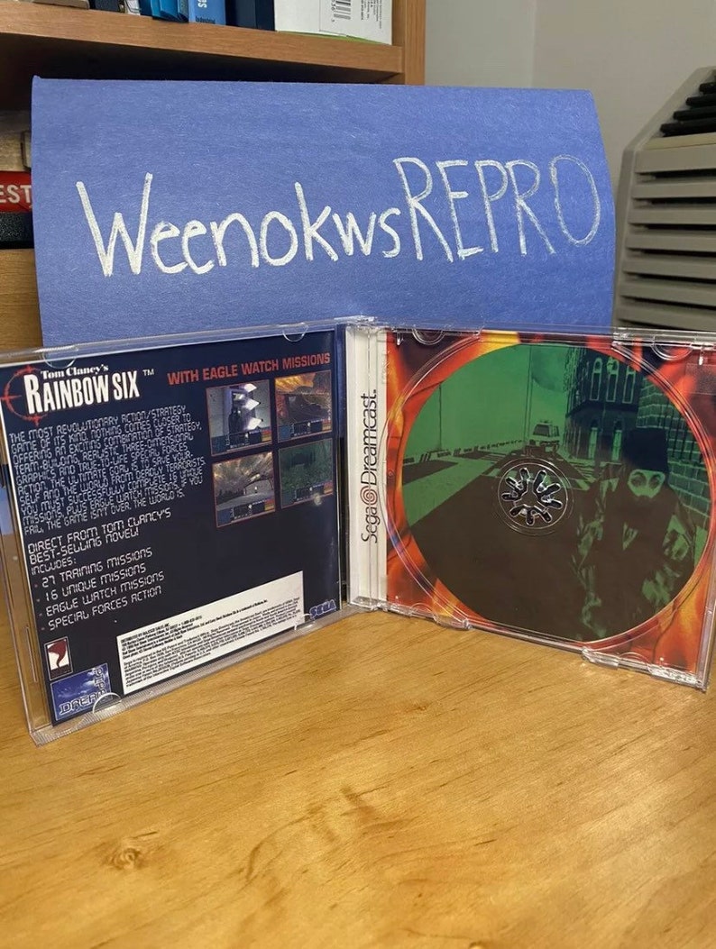 May include: A Sega Dreamcast game case for Tom Clancy's Rainbow Six. The front of the case shows a dark, urban scene with a person in the foreground. The back of the case has a white background with black text and a picture of the game's logo. The text on the back of the case reads "With Eagle Watch Missions".