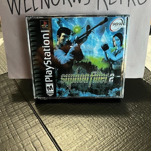 May include: A PlayStation game case for Syphon Filter 2, featuring a blue and green background with two characters holding weapons. The case is labeled with a "Mature" rating and the 989 Studios logo.