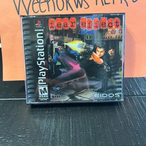 Può includere: Una custodia per gioco PlayStation per il gioco "Fear Effect" con sfondo nero e una "M" rossa per la classificazione "Mature". Il gioco presenta una donna con un abito nero che tiene una pistola e un uomo con una camicia blu che tiene una pistola. Il gioco è classificato "M" per "Mature" dal PEGI.