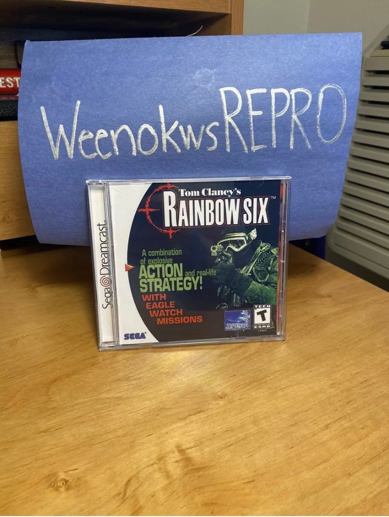 May include: A Sega Dreamcast game case for Tom Clancy's Rainbow Six. The case is green, black, and white with a picture of a soldier wearing a helmet and holding a gun. The text on the case reads "Tom Clancy's Rainbow Six" and "A combination of explosive action and real-life strategy!"