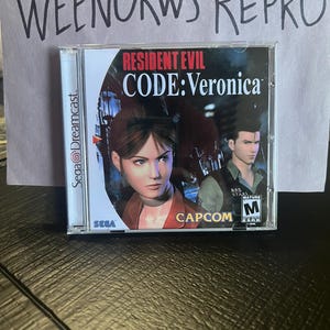 Könnte beinhalten: Ein Sega Dreamcast-Spielecase für Resident Evil: Code Veronica. Das Case ist silberfarben mit einem schwarzen und roten Design. Das Spiel zeigt eine Frau mit braunen Haaren und einem roten Shirt sowie einen Mann mit braunen Haaren und einem grünen Shirt. Das Spiel ist von der USK mit M für Erwachsene freigegeben.